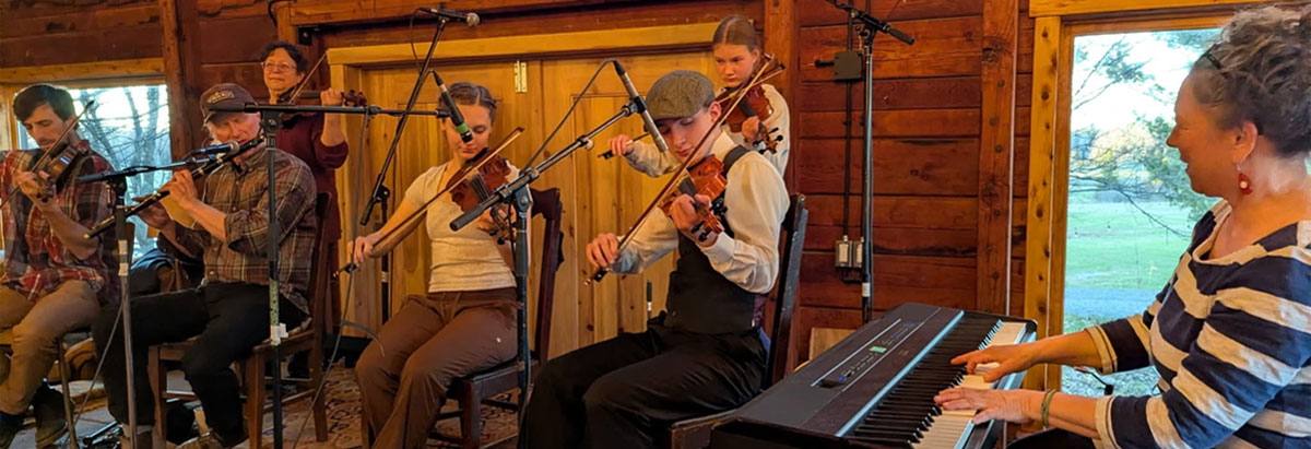 Tab 1: A group of seated musicians perform against a rustic cabin wall.