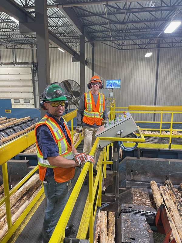 Two men in safety vests and helmets feed logs into a sawmill.