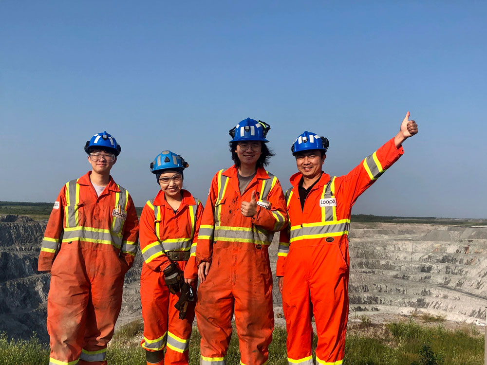Four people dressed in orange workwear standing outside of an open mine. One person has their hand in the air.