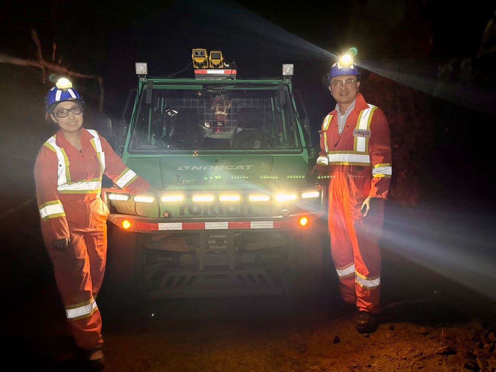 Two people stand in front of an all-terrain vehicle with headlights illuminated in an underground mine.