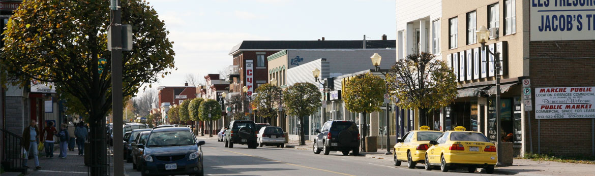 Car-lined street and downtown section of a community featuring shops, businesses and pedestrians on the sidewalk