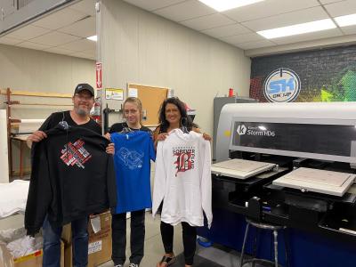 Three people stand in front of the new printing equipment showing off newly printed apparel in their hands